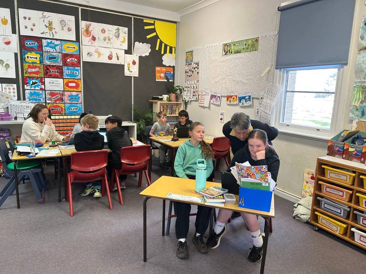 A classroom with children sitting at desks learning. One desk has a teacher sitting with the children helping them work. Another desk has another teacher leaning over the back of the students assisting them with their work. It is a bright classroom with plenty of artwork around the room.