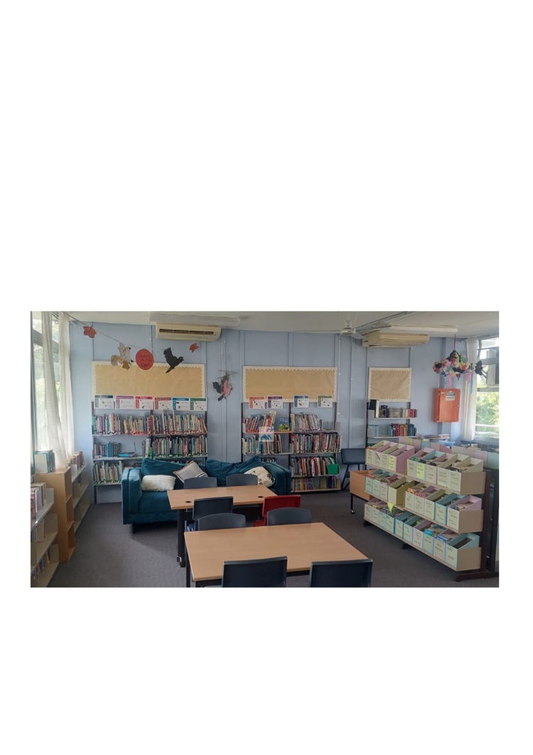 A bright and light library room with 2 tables in the centre of the room with chairs around them. Behind that is a comfortable blue lounge with cushions on it and all around the room are book shelves full of books. There is also some artwork hanging from the ceiling.