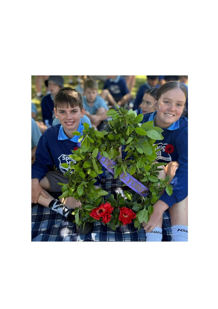 Two year 6 students, one boy and one girl sitting on a mat holding a wreath in front of them. The wreath is full of green leaves and has 2 large red ANZAC poppies at the bottom of it. It also has a purple banner diagonally across it stating