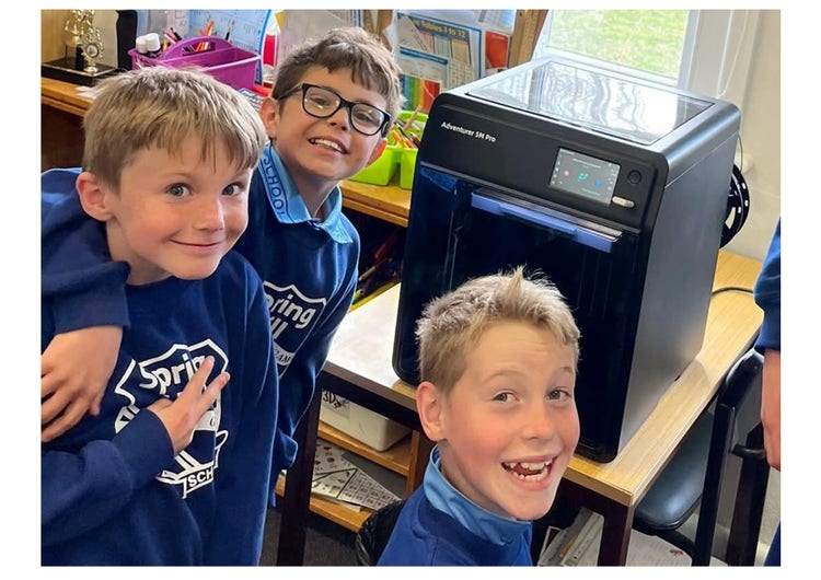 Three children smiling at the camera with a 3D printer in the background.