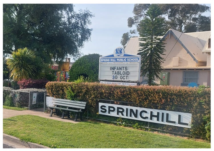 A photo of the front of the school taken from the road. There is a wire fence with a hedge growing all along the back of it and in the fence is a big white sign with black writing stating