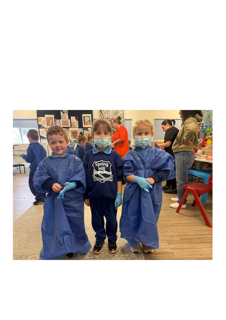 Three K-1 students looking at the camera. The first child is wearing a gown and gloves and is smiling at the camera. The second child is wearing a medical face mask and gloves and the third child is wearing gloves, mask and gown. There are students and teachers in the background and they are in the art room..