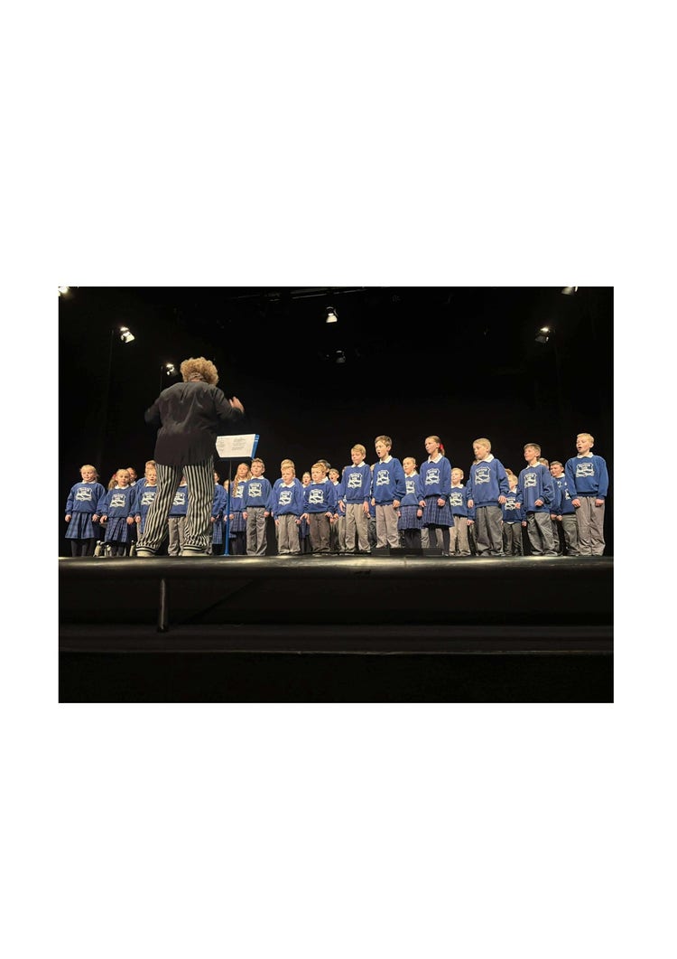 A photo of the whole school on a stage with a black background. They are participating in verse speaking and are all standing in position. Mrs Mac is out the front with her back to the camera facing the children instructing them. She is wearing a black shirt and black and white pants and the children are watching her for direction.