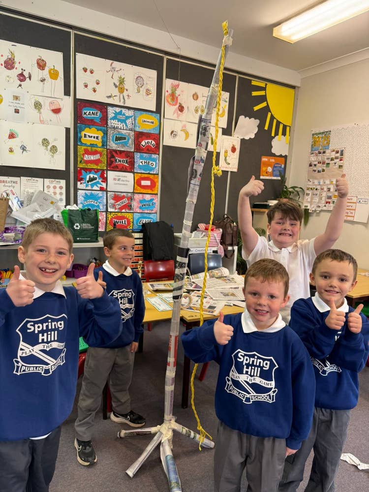 A photo of five students in the K-1 classroom with big smiles and giving a thumbs up to the camera. They have built a large paper tower, over 6 feet tall) with a yellow rope hanging from the top of it.