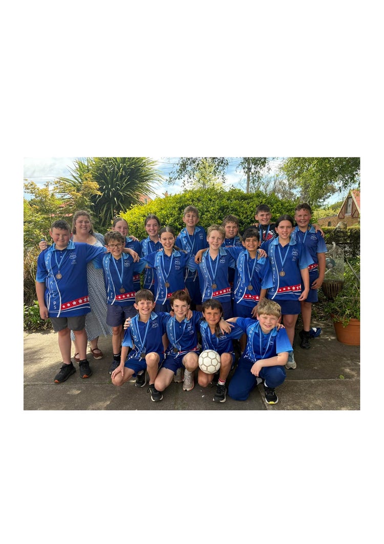 A full group photo of both female and male students with their teacher, Miss Em. There are students standing at the back and the students crouched down at the front are holding a soccer ball. They are standing in the playground in front of the green shrubs at the front of the school. All students are smiling brightly and have their arms around each other.