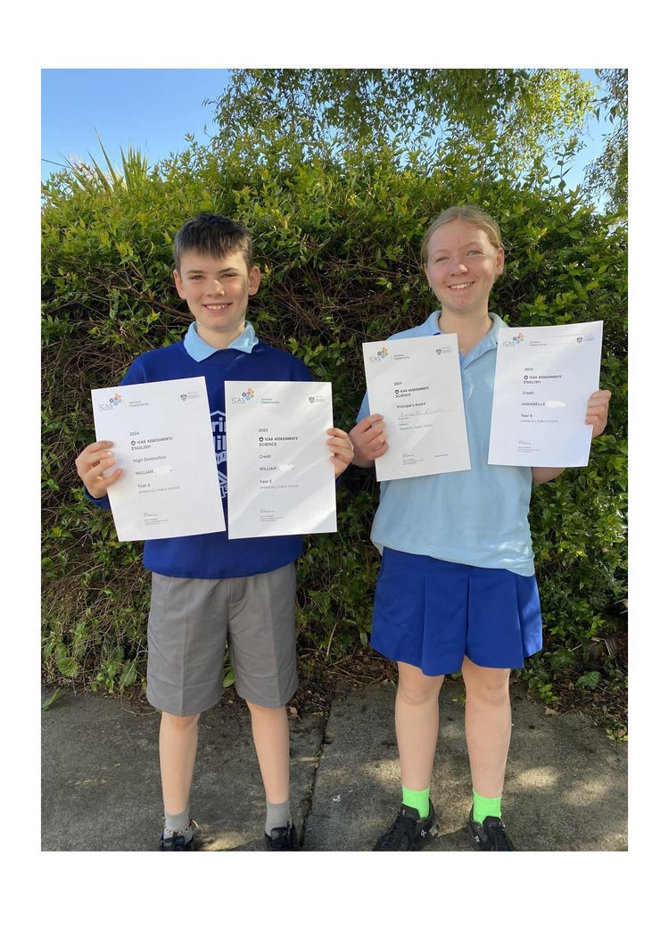 Two students, a boy and a girl, smiling at the camera holding up two certificates each. They are standing in front of the bushes at the front of the school.