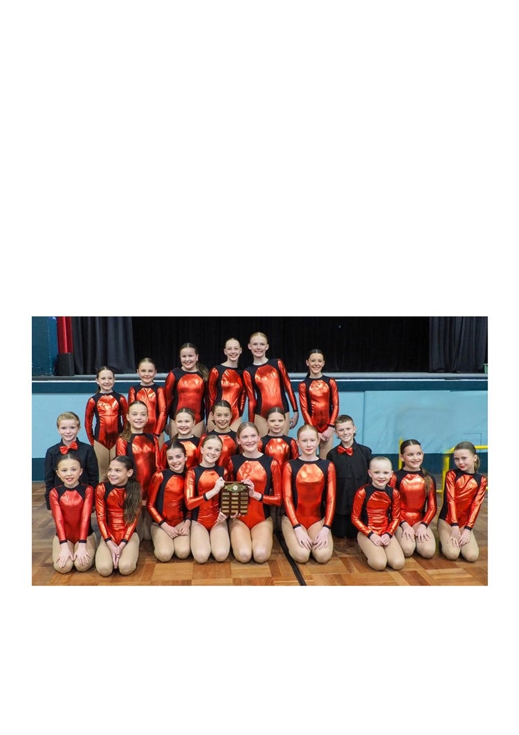 A group photo of the Orange Small Schools Dance Troupe. The girls are wearing red and black leotards and the boys are wearing black trousers and long sleeve shirts with red ties. They are all sitting and standing in a posed photo smiling at the camera in front of the stage at the Orange Function Centre.